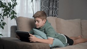 Happy teenage boy using tablet on sofa. Scrolling social media, surfing internet, enjoying time at home alone. Smiling child laying on coach in living room with device. Listen to music, watching video