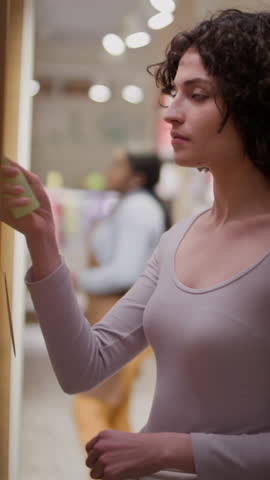 Medium vertical shot of Caucasian girl with curly brown hair standing in beauty shop, picking up tester bottle of perfume or skin mist, spraying on wrist, smelling, nodding and smiling