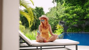Redhead woman in summer dress relaxes by poolside, sips cocktail. Enjoys tropical lifestyle, leisure in luxury setting. Makes video call on smartphone, laughs, chats, connects with friends. Slowmo - Powered by Shutterstock - Get 15% off with code: PIKWIZARD15