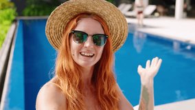 Redhead woman with sunglasses, straw enjoys poolside leisure, sips cocktail, waves in friendly video call. Relaxed, sunny resort atmosphere. Media, tech savvy millennial moment on smartphone. Slowmo - Powered by Shutterstock - Get 15% off with code: PIKWIZARD15