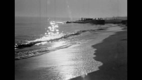 Sunset on sea coast on black white grainy film archive. Sun rays shine in calm, slow sea waves arriving on sandy shore. Archival footage of beautiful seascape. Vintage ocean shore nature. Retro 1980s
