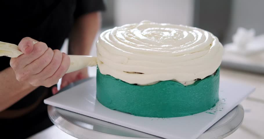Close-up of the hands of a young female pastry chef applying white buttercream to the surface of a cake on a turntable. The process of work in production in a confectionery shop. Cake decoration.