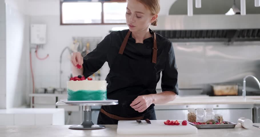 Medium shot view of a bright production in a confectionery shop. A young red haired charming woman pastry chef concentrates on decorating a cake with fresh berries. Professional work process. Royalty-Free Stock Footage #3467727349