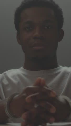 Vertical zoom out portrait of arrested Black man looking frustratedly at camera sitting at desk in interrogation room with his hands locked in handcuffs Royalty-Free Stock Footage #3467734185