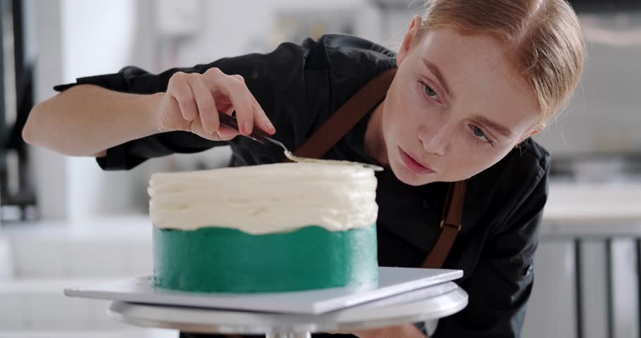 Portrait of a young red haired female pastry chef who looks at the cake with concentration and distributes the cream in an even layer over its surface. The process of preparing in a production kitchen Royalty-Free Stock Footage #3467735903