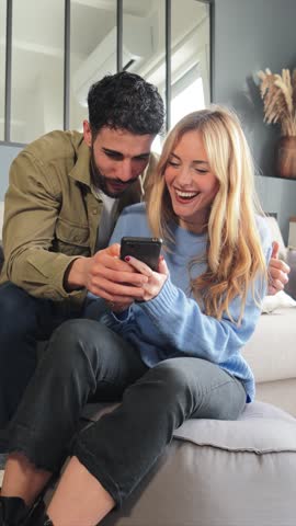 Vertical. Young caucasian couple using a cellphone to do shopping online. Man and woman watching the social media with a smartphone. Adult friends having fun browsing on internet with a mobile phone