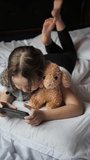 An eight-year-old girl lying on her stomach in bed hugging soft toys uses a smartphone to play online. A child has fun with plush animals and a mobile phone, dangling his legs. Hobbies and leisure