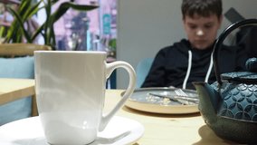 boy is sitting in cafe drinking tea, with a mobile phone and watching video or read article. teenager plays online on the phone, types a message to friends, communicates on social networks.