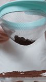 A close-up shot from below of a woman wearing a white apron, sprinkling cocoa powder on a tiramisu cake. slow motion. Vertical video.