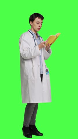 Side view Medic specialist reading a healthcare literature book to develop her medical expertise in the industry, stands against greenscreen backdrop. Clever doctor in white coat enjoys lecture Royalty-Free Stock Footage #3467886675
