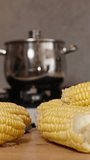 Vertical video. In the foreground, corn is placed on the table as a woman adds them into a pot of boiling water.