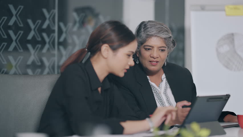 Indian business women sitting on chair using tablet look screen discuss new project planning share ideas indoor job office. Young adult colleagues old elder female doing work task together workplace Royalty-Free Stock Footage #3468089143
