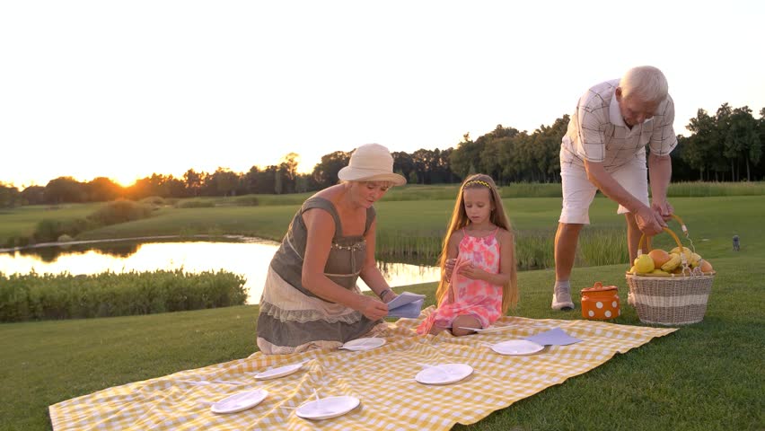 Elderly people and child on picnic. Old man placing basket with fruits on picnic cloth, summer nature. People and picnic cloth.