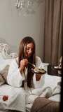 Two young women in bathrobes are having breakfast in bed. Healthy breakfast cheesecakes. Hotel holiday concept. Vertical video