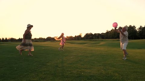 Child and grandparents playing ball. Couple of senior people and their granddaughter having fun on green meadow at sunset, slow motion. Family weeked on nature.