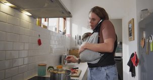 Video of caucasian mother with newborn baby in baby carrier using smartphone in kitchen. motherhood, parental love and taking care of newborn baby concept digitally generated video.