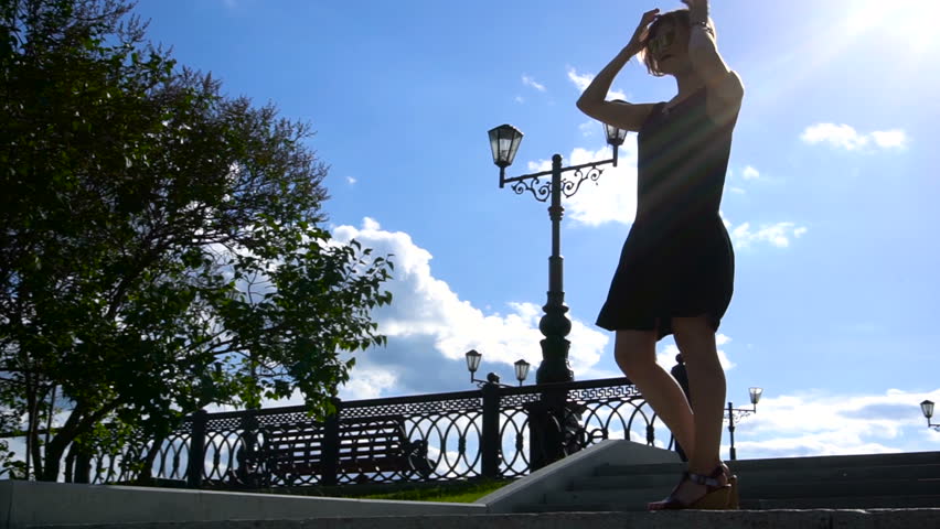 Silhouette of romantic woman wear black short dress on top park staircase in the wind. windy weather. Beautiful young woman against sun background