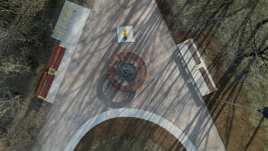 circle-shaped wood bench with a tree growing in the middle. swings and sandy paths in the park. a bed of flowering bulbs. yellow gates and pine trees looking vertically down, drinking fountain