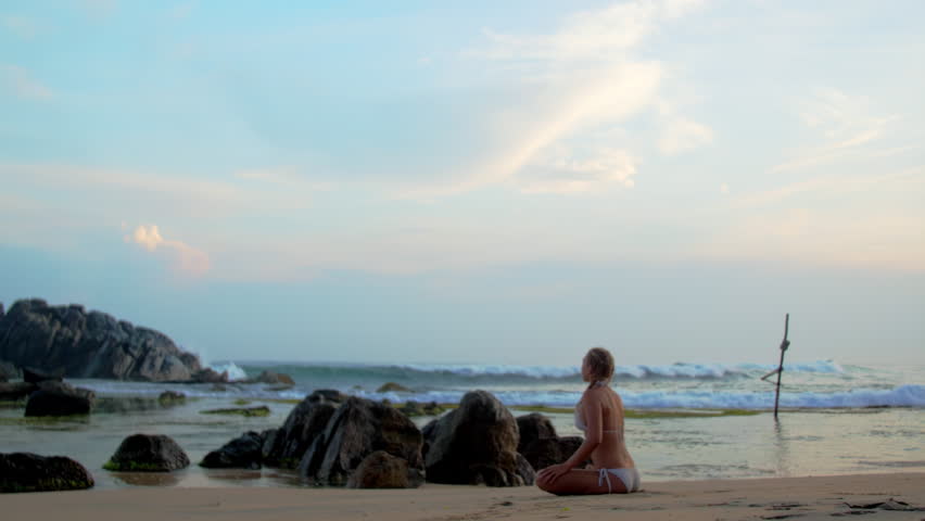 Woman in bikini meditates sitting in lotus pose near rocks on beach by ocean. Lady with braids finds inner peace at sunset