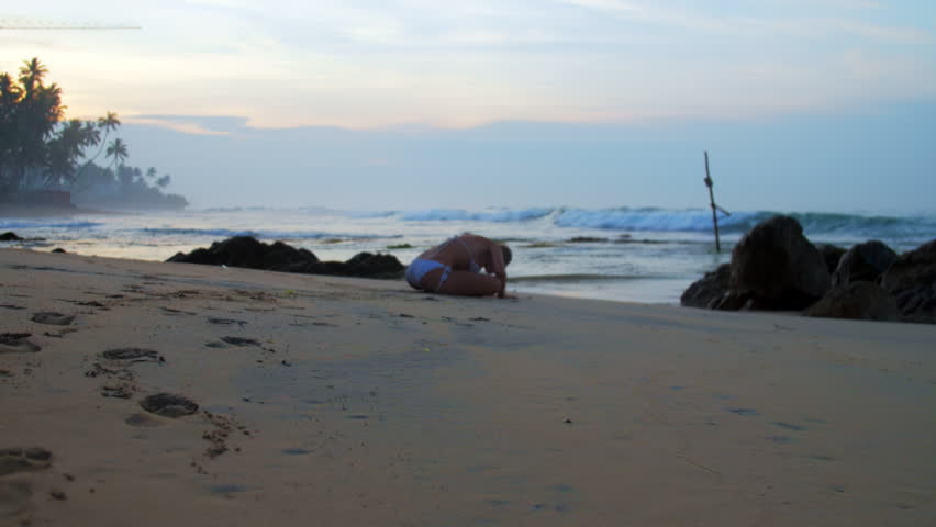 Silhouette of young woman doing yoga asanas on sand beach by waves at sunset. Woman stretching on tropical sand beach by ocean slow motion