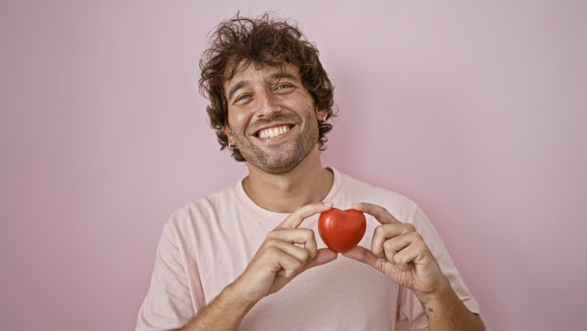 Smiling young man holding a red heart against a pink background, depicting positivity and love