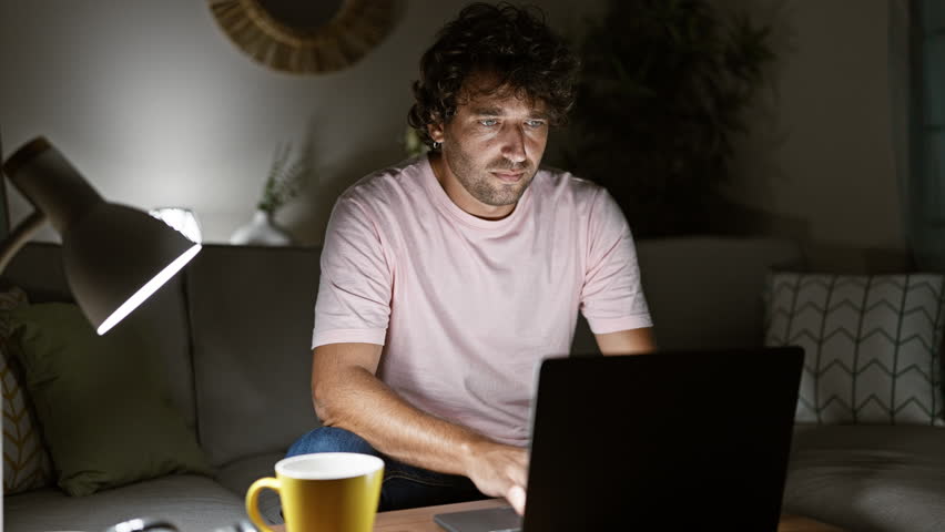 Tired man using laptop at night in living room, stressed male with coffee, freelance work