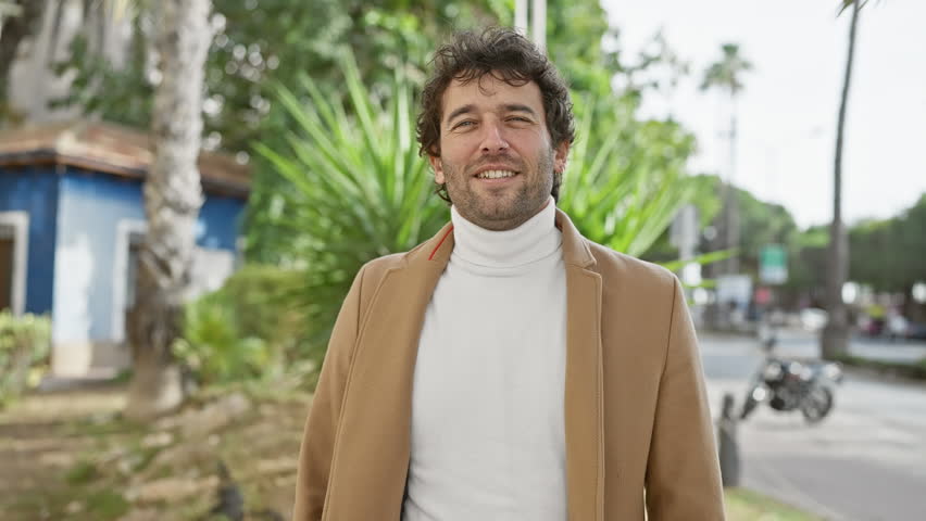 A smiling young hispanic man in a coat standing on a city street extends his hand towards the camera, giving a sense of welcoming and friendliness.