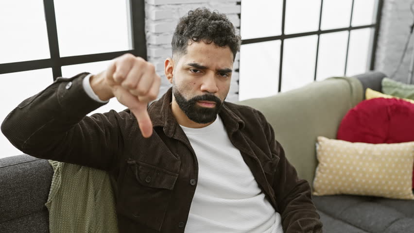 Unhappy young man at home showing his angry expression, thumbs down gesture in rejection of negative and bad