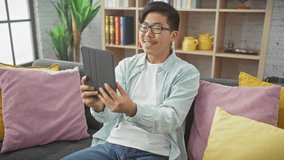 Young asian man enjoying a video call at home, smiling in a cozy living room setting.