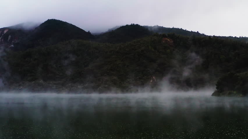 Static aerial shot of steam rising over geothermal lake in New Zealand. Surrounded by forest and mountains.