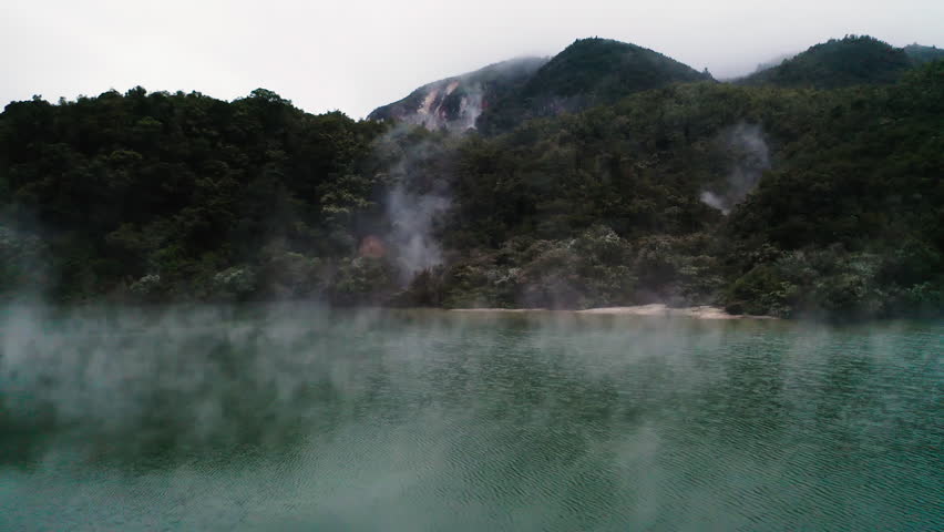 Fly through steam rising over geothermal lake and lush forest in New Zealand, North Island. Aerial drone fly over.