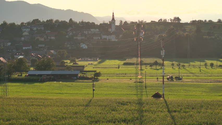 AERIAL: A slow motion video flying above a neatly cultivated piece of land. Two farmers walking in front of a driving tractor. A small sunlit village with a picturesque church rises in the background.