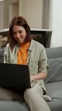 Vertical video Happy brunette woman in a light green and orange T-shirt sits on a gray sofa and looks at something in her gray laptop in a modern apartment