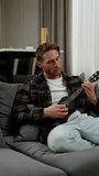 Vertical video Happy blond man with stubble in a plaid shirt and white T-shirt sits on a gray sofa and plays a black ukulele in a modern apartment during the day
