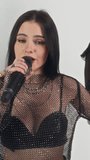 Vertical chest up footage of attractive Caucasian vocalist of glam rock band wearing mesh top and singing into microphone while performing song in studio on white background and looking at camera