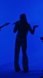 Vertical full footage of silhouettes of musical trio of female singer and musicians gigging in professional studio with blue colored neon filter
