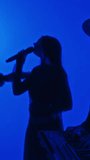 Vertical side footage of silhouettes of rock band of keyboarder, guitarist and singer performing music in obscure professional studio with blue neon filter