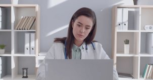 Portrait of focused concentrated professional young female medical doctor in white uniform with stethoscope working at the desk in office on laptop. Physician woman working in clinic. 4k video.