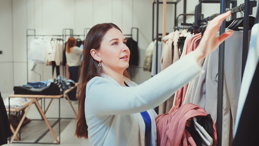 Young woman in chic blazer enthusiastically browses through variety of clothes in fashion store. Spirit of shopaholic