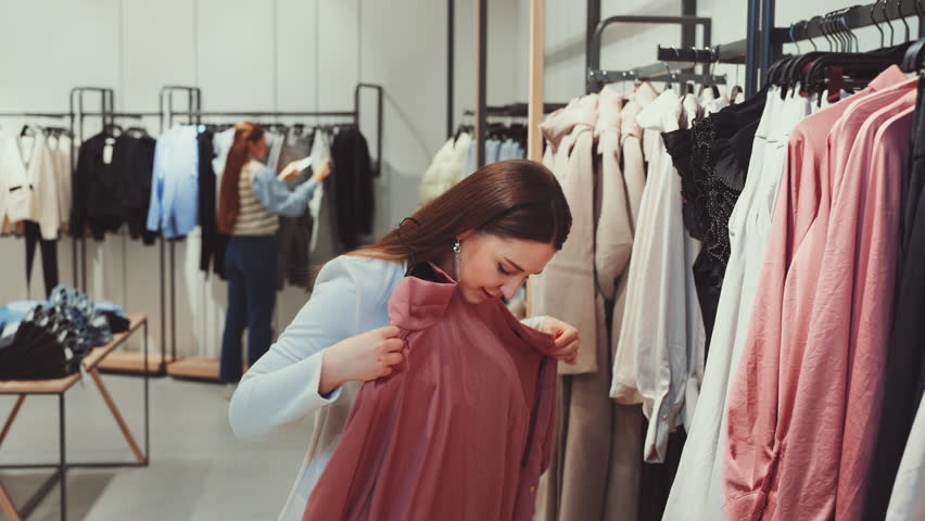 Woman in clothing store smiles while holding up and looking at terracotta dress, with other clothing items and customer in background