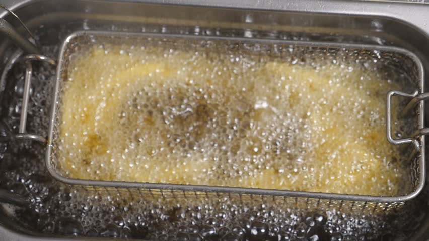 Top view of male cook shaking a lattice with french fries being prepared in hot boiling oil. Chef lowering a grid with potatoes into deep fryer. Process of making fast food at cuisine. Slow motion