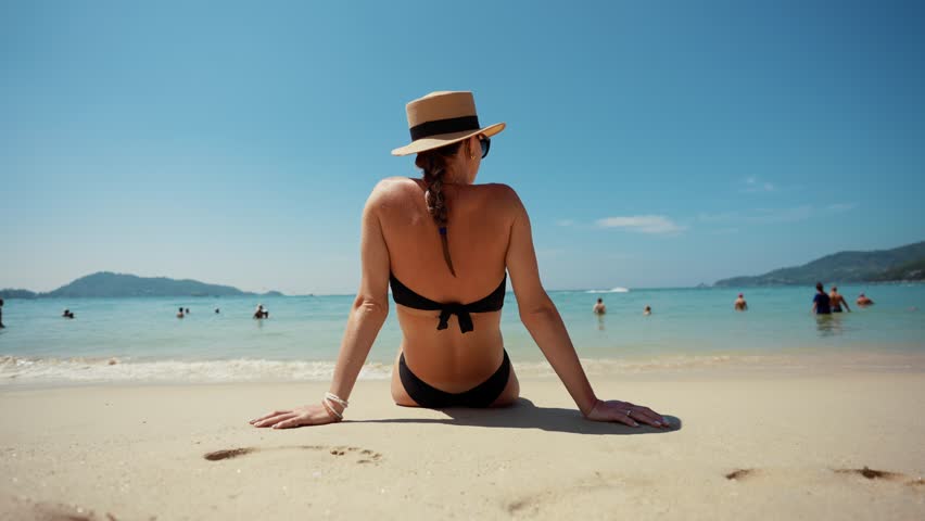 Blonde woman in black bikini straw hat. Girl sitting on ocean beach in tropical paradise island with water waves, admire white sand in sunny weather back view. Beach resting, travel trip, tourism.