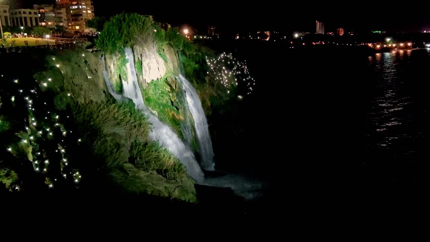 Nght Waterfall with Beautiful Lighting Flowing into Sea from Great Height of Steep Shore, against Background of Night City Lights., Concept of Merging City with Nature.