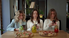 Three women at a table with tableware, pizzas, and drinks, looking at phones
