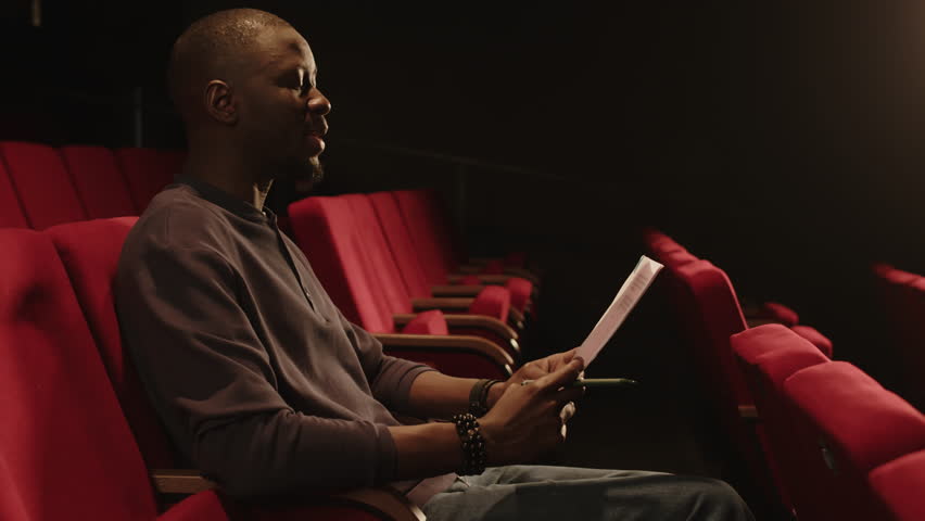 Medium shot of young expressive Black male actor sitting on red seat and revising monologue during auditions in theater Royalty-Free Stock Footage #3468947805