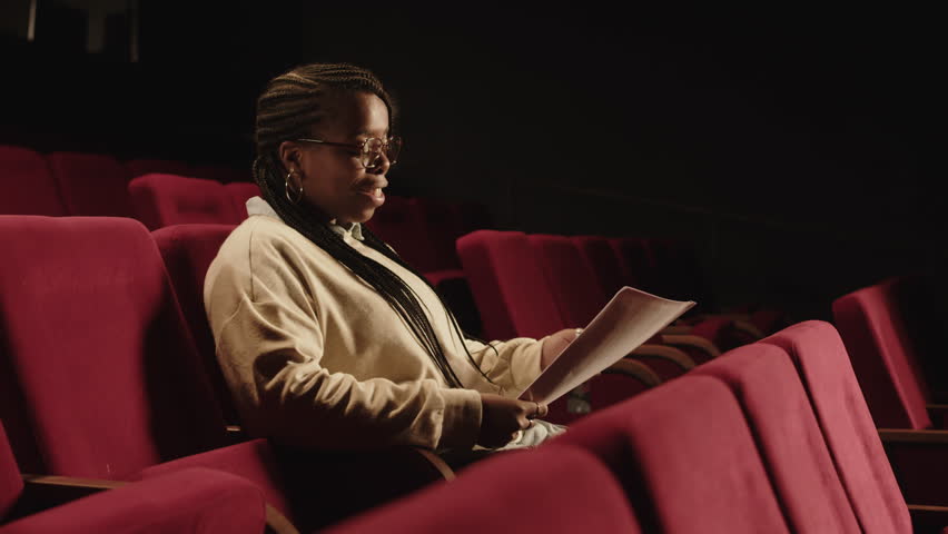 Side view medium shot of young Black theatre actress sitting on red seat in dark theatre auditorium reading comedy script and laughing while preparing for auditions Royalty-Free Stock Footage #3468961789