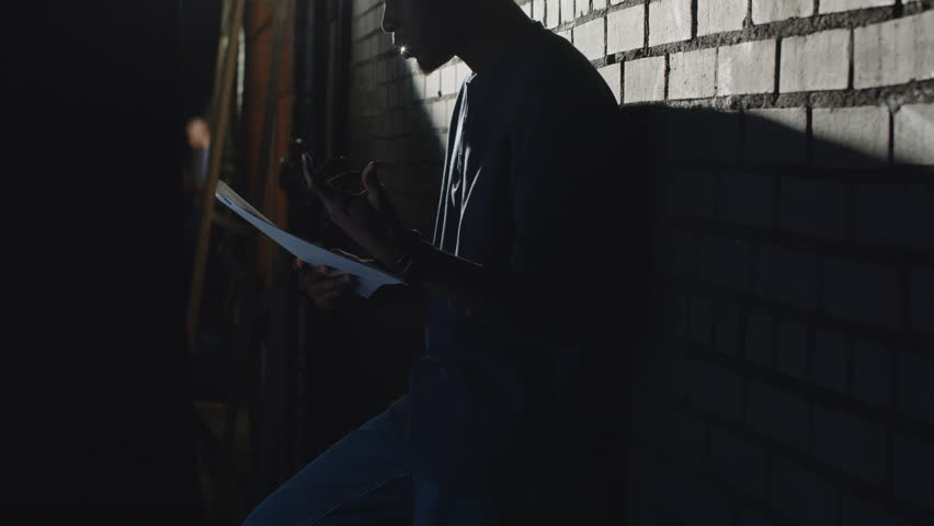 Lowkey medium shot of young Black male standup comedian rehearsing his monologue and laughing while standing in dark against brick wall corridor before going on stage Royalty-Free Stock Footage #3468964573