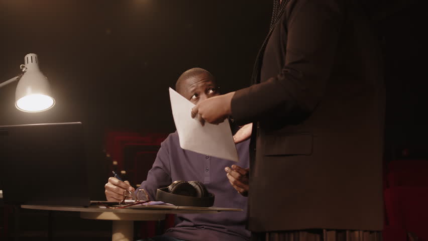 Medium shot of young Black man and his female co-author discussing standup jokes and concert program while rehearsing in low light amateur theater Royalty-Free Stock Footage #3468986595