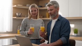 Happy adult middle-aged family at home kitchen using laptop discuss talking computer online booking order food delivery man close pc talk to woman finish work couple drinking healthy fresh fruit juice - Powered by Shutterstock - Get 15% off with code: PIKWIZARD15