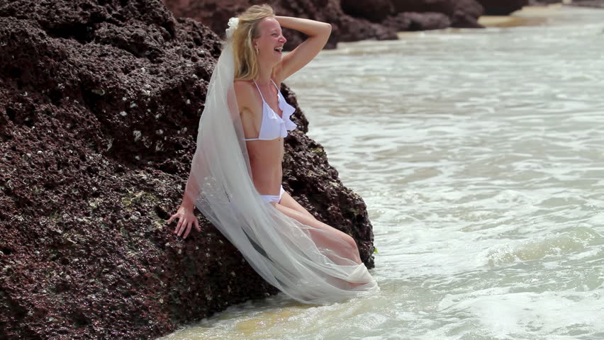 Cheerful bride in bikini with long veil leans on rock basking in ocean waves. Blonde bride laughs posing for memorable photo on wedding day